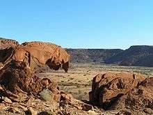 A sandstone rock resembling the mouth of a lion. In the distance a patch of desert savanna is flanked by a table mountain.