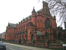 A red brick church with a rose window, a bellcote and a series of gabled buttresses