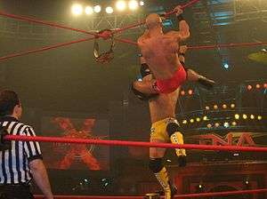 Two men, one wearing red trunks and the other wearing yellow trunks, hanging from red crossed ropes that form an "x" above a wrestling ring as a referee watches.