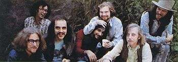 Seven long-haired young men in casual clothes sit close together, look forward, and smile. One of the men in the middle of the shot leans on another man's head while holding a cup in his other hand.