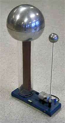 Large metal sphere supported on a clear plastic column, inside of which a rubber belt can be seen. A smaller sphere is supported on a metal rod. Both are mounted to a baseplate, on which there is a small driving electric motor.