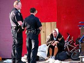 Two VPD officers in short sleeves stand talking to a woman sitting on the sidewalk, wearing in a heavy coat