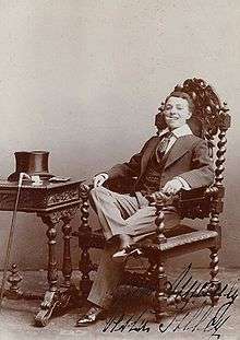 Sepia-tone image of a woman dressed in a man's suit, tie, and waistcoat and seated in a large armchair next to a table on which a top hat sits and cane rests nearby. Her collar is overlarge and her shoes dainty but patent leather, spats, and 2-inch heels. She is grinning at the viewer with a cigarette clenched in her teeth.