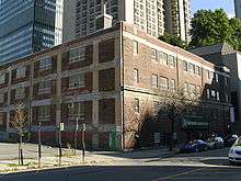 A three-storey brown-bricked building. It has numerous windows and the concrete of each floor is exposed to the outside. Behind it are several modern tall buildings.