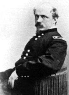 Head and torso of a balding white man with a Van Dyke beard and mustache, sitting in a chair with his left arm lying on the arm rest. He is wearing a dark double-breasted military jacket.