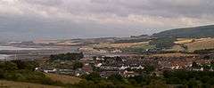 View of multiple houses with sea on the left and hills in the background.