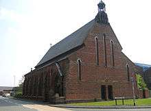 A plain brick church seen mainly from the end, with a steeply pitched roof, a doorway and buttresses on the left side, and a small bellcote with one bell on the gable end