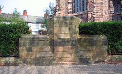 A sandstone structure in the shape of an inverted T with an inscription in Gothic script along the top; behind is part of the church to the right and a row of houses to the left