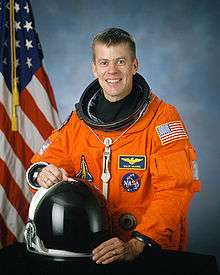 man in space suit holding helmet, American flag in background