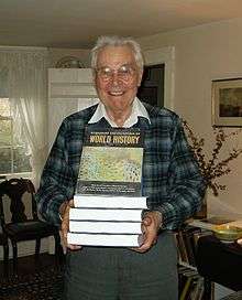 Smiling older man holding a stack of books in front of him; the top one is tilted up so the title, World History, is visible.