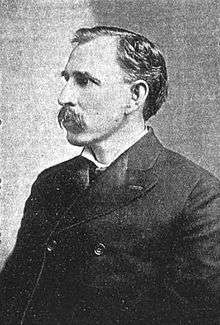 A mustachioed man in his mid-forties with black hair. He is wearing a black coat buttoned at the high chest and facing left