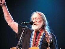 A Caucasian male singing into a microphone. The male has a white beard and long white braided hair that is on his left shoulder and has his right arm raised in the air.