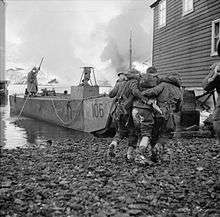 two men each side of a wounded man, helping him towards a boat docked between houses