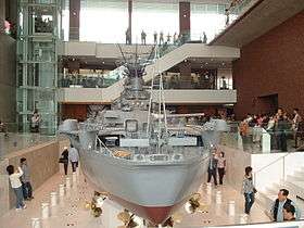 Rear view of a very large model of a battleship in an open gallery. It is surrounded by people examining the model and taking photographs of it