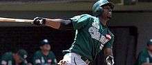 A dark-skinned man wearing a green baseball jersey and cap swinging a baseball bat