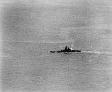 A large area of ocean with a warship in the middle distance. A plume of smoke is coming from the rear of the ship's superstructure, and the ship appears to be leaning to the left.