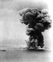 A view of the ocean stretching to the horizon with the silhouette of a distant small warship visible to the left. To the right an enormous mushroom cloud rises high into the sky.