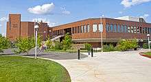 A modern irregularly shaped rectilinear brick building with concrete walkways curving up to it through grass and small trees, with two people sitting on a bench in front. A sign in front and vertical lettering on the building read "Yellowknife City Hall".