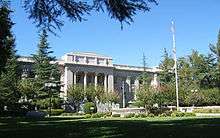 A large white stone governmental building, with pillars in front.  Parts of the facade are obscured by evergreen trees.