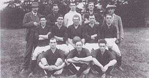 A faded photograph of a posed group of men, outdoors. At the front are three men seated on the ground, each dressed in sports clothing, a dark-coloured shirt, white shorts, dark socks and boots. In the middle row five men are sitting on benches, each dressed in sports clothing. Standing at the back are two men, weating hats and coats, and three men in sports clothing.