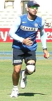 A man in a dark blue cap wearing green glasses, along with a light blue shirt with the words "SAHARA" on the front and dark blue or navy shorts.