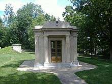 A photograph of a grey-bricked building with golden double doors under an engraving reading "ZACHARY TAYLOR" all under a cloudy, blue sky