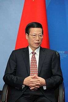 a seated man with a short haircut, wearing glasses, a white shirt, a suit and a red tie with white dots